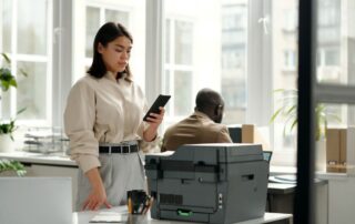 A woman in an office using a smartphone next to a business copier, highlighting the copier's role in supporting daily office tasks and enhancing productivity.