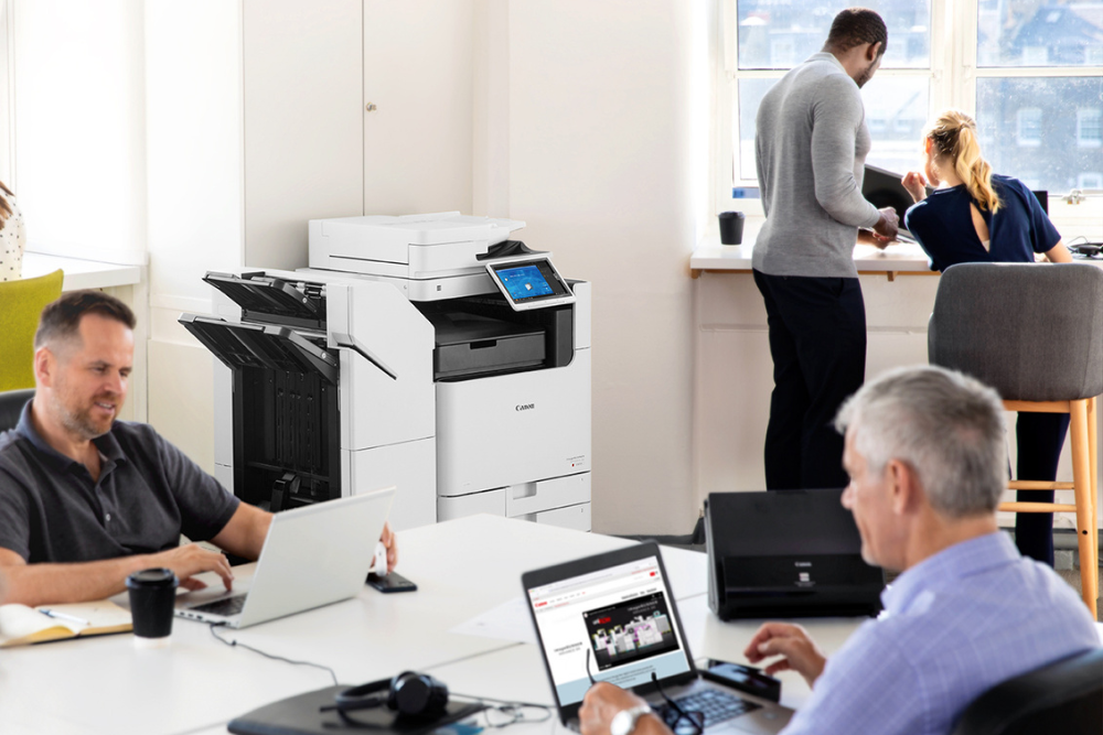 Office setting with employees working on laptops and a multifunction printer in the background. The multifunction printer is prominently featured, highlighting its role in the busy office environment.