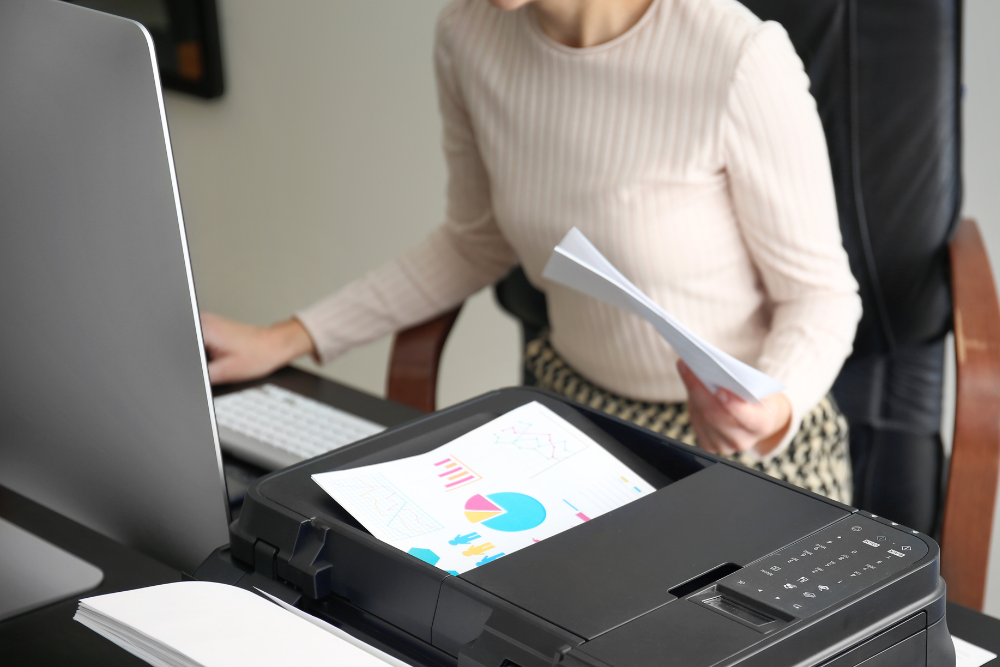 A woman working at her desk with documents and a colorful chart being printed from a nearby printer. This image illustrates the efficiency of Managed Print Services (MPS), showcasing how businesses can streamline their printing processes and manage documents effectively.