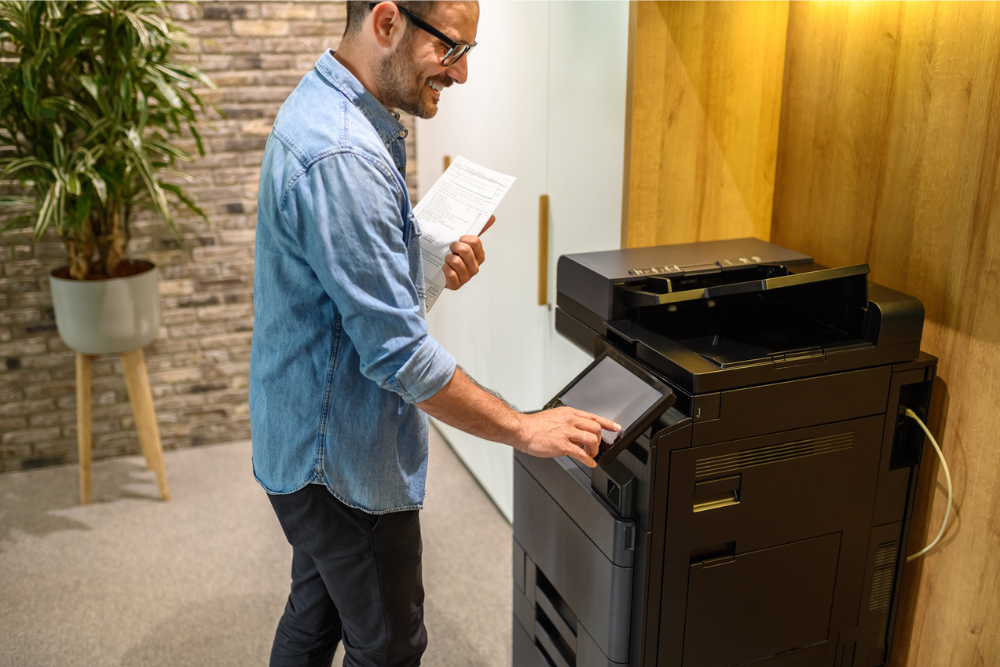 A man using a color MFP (multifunction printer) in a modern office, interacting with the touchscreen interface while holding a document, highlighting the convenience of advanced printing technology.